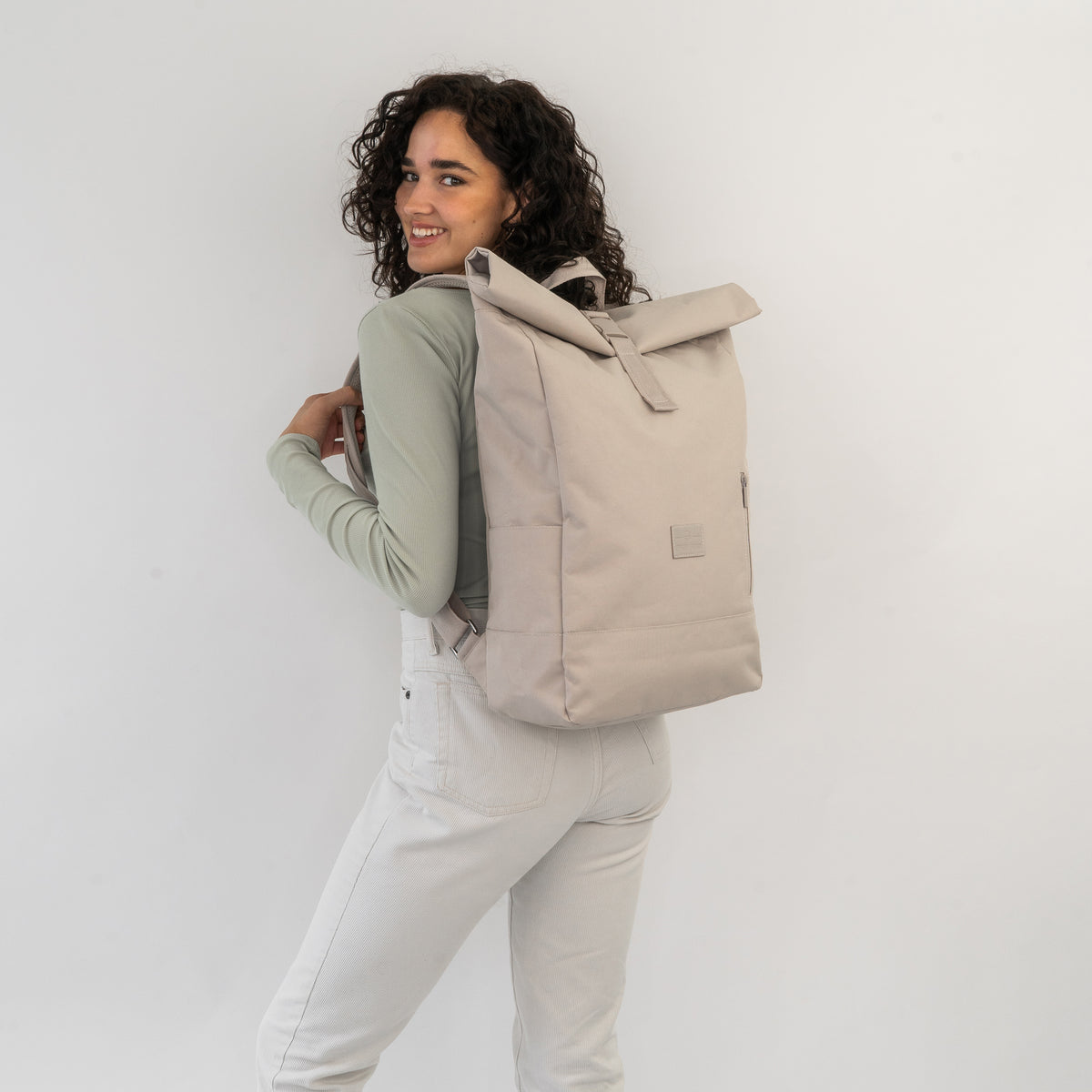Frau mit lockigem dunklem Haar trägt den hellbeigen Rolltop Rucksack 'Robin Large' von Johnny Urban auf dem Rücken