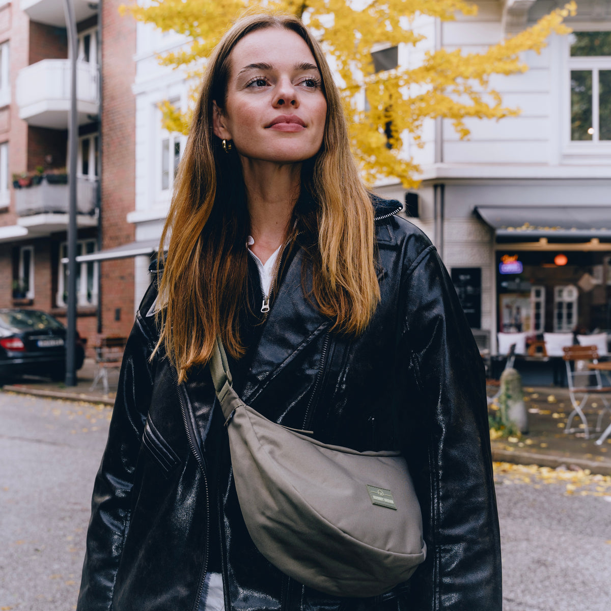 Frau mit langen blonden Haaren trägt schwarze Lederjacke und graue Half Moon Bag 'Ally Medium' quer über die Schulter in urbaner Herbstumgebung