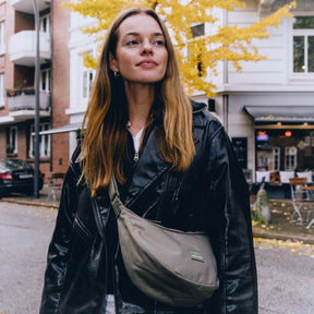 Frau mit langen blonden Haaren trägt schwarze Lederjacke und graue Half Moon Bag 'Ally Medium' quer über die Schulter in urbaner Herbstumgebung