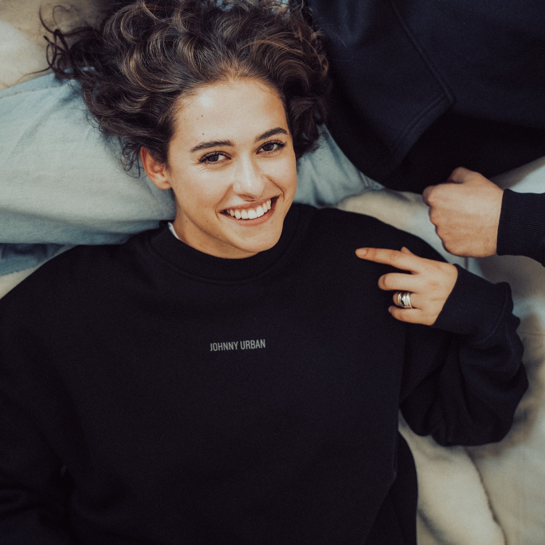 Frau liegt lachend auf dem Rücken, trägt schwarzen Oversized-Crewneck mit 'Johnny Urban' Print