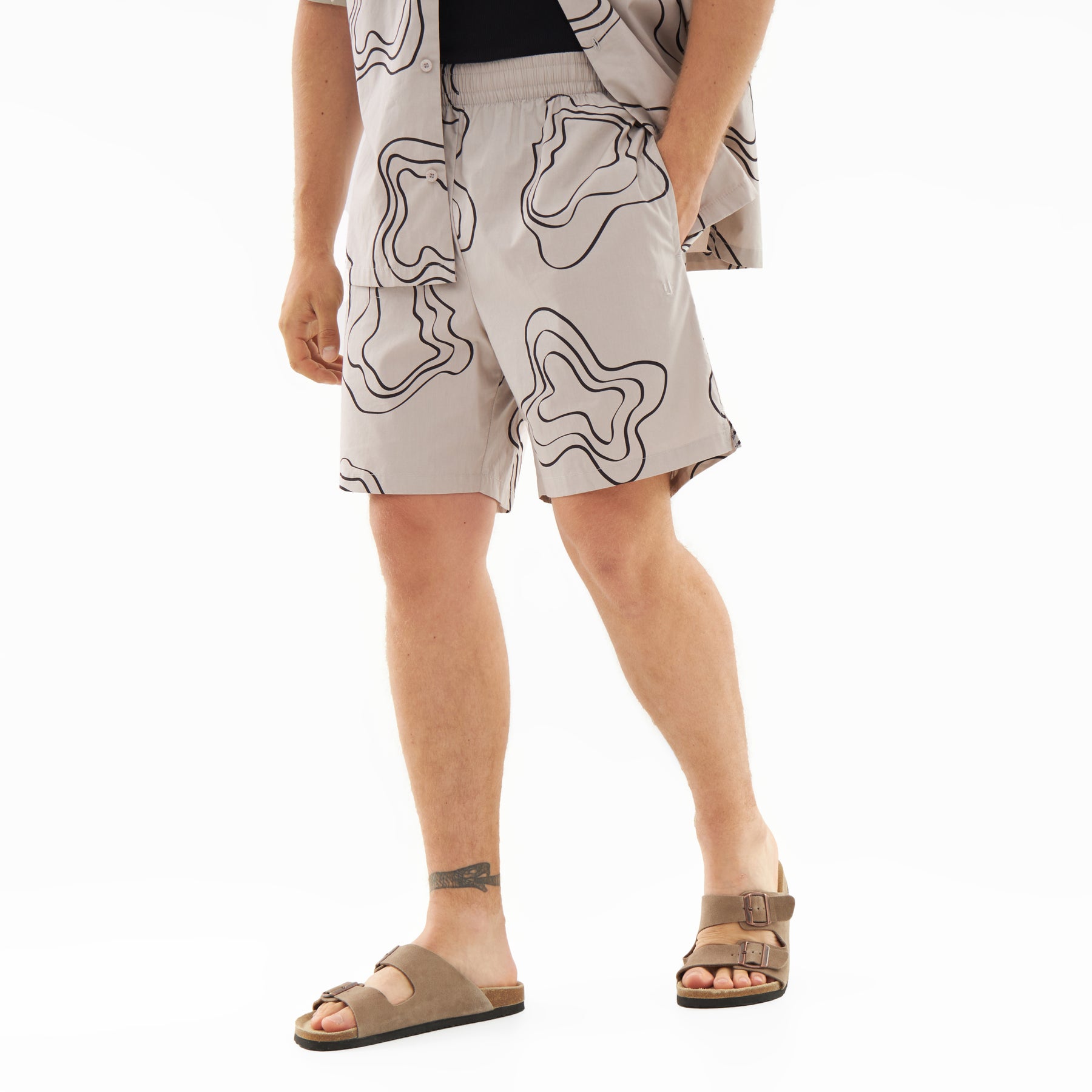 Mann trägt beiges Shorts-Set 'Spencer' mit schwarzen abstrakten Linienmuster und passende Sandalen