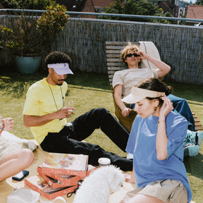 Drei Personen entspannen im Garten bei Sonnenlicht, zwei tragen Sandfarbene Sun Visors 'Terry', eine trinkt aus einem Glas