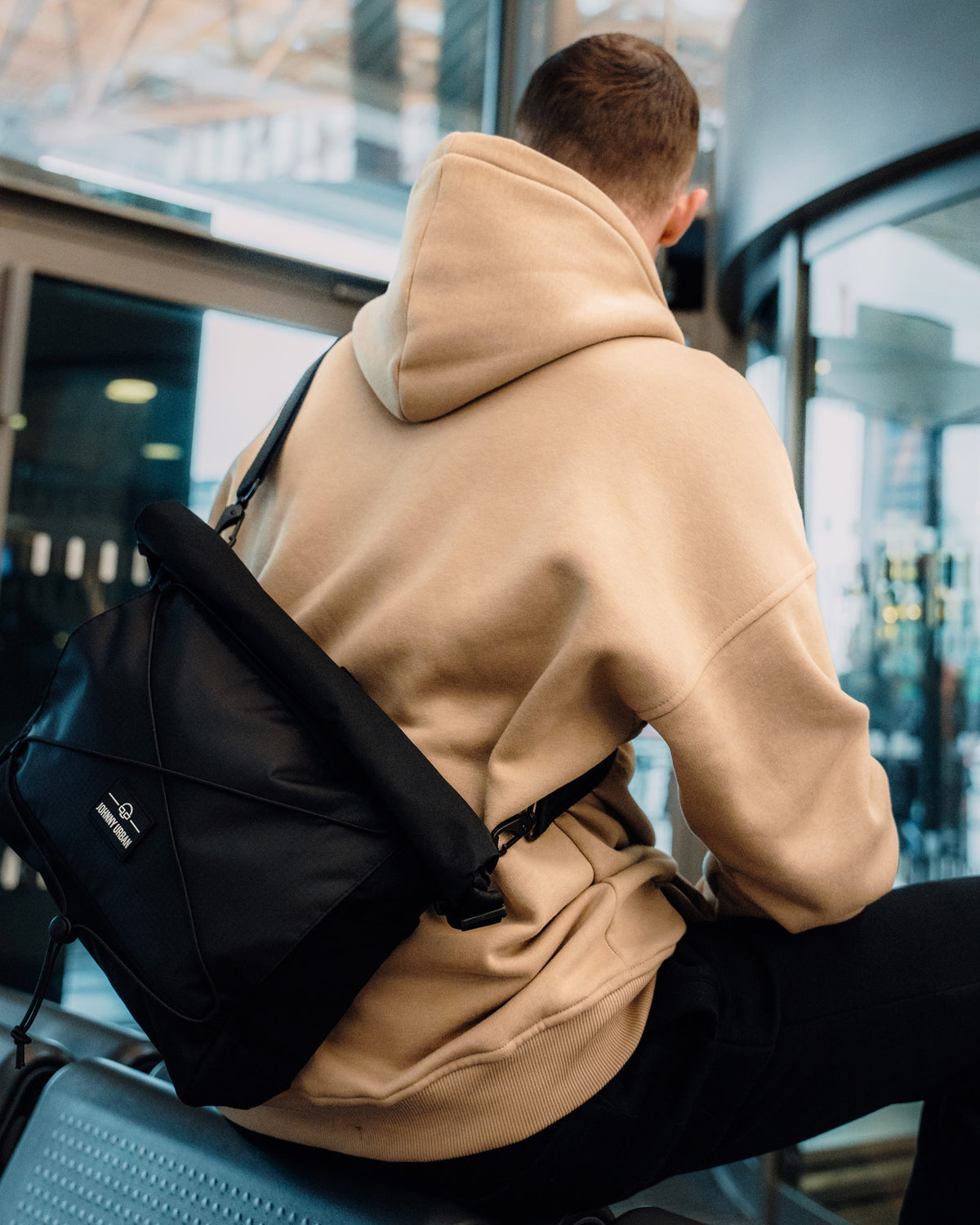 Schwarze Johnny Urban Umhängetasche 'Dylan' aus robustem Material, getragen von einem Mann im beigen Hoodie in moderner Umgebung