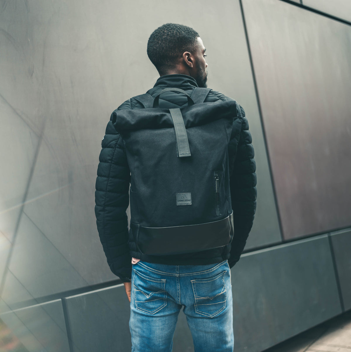 Mann trägt schwarzen Rolltop Rucksack 'Adam Large' von Johnny Urban, sieht nach vorn an Wand aus