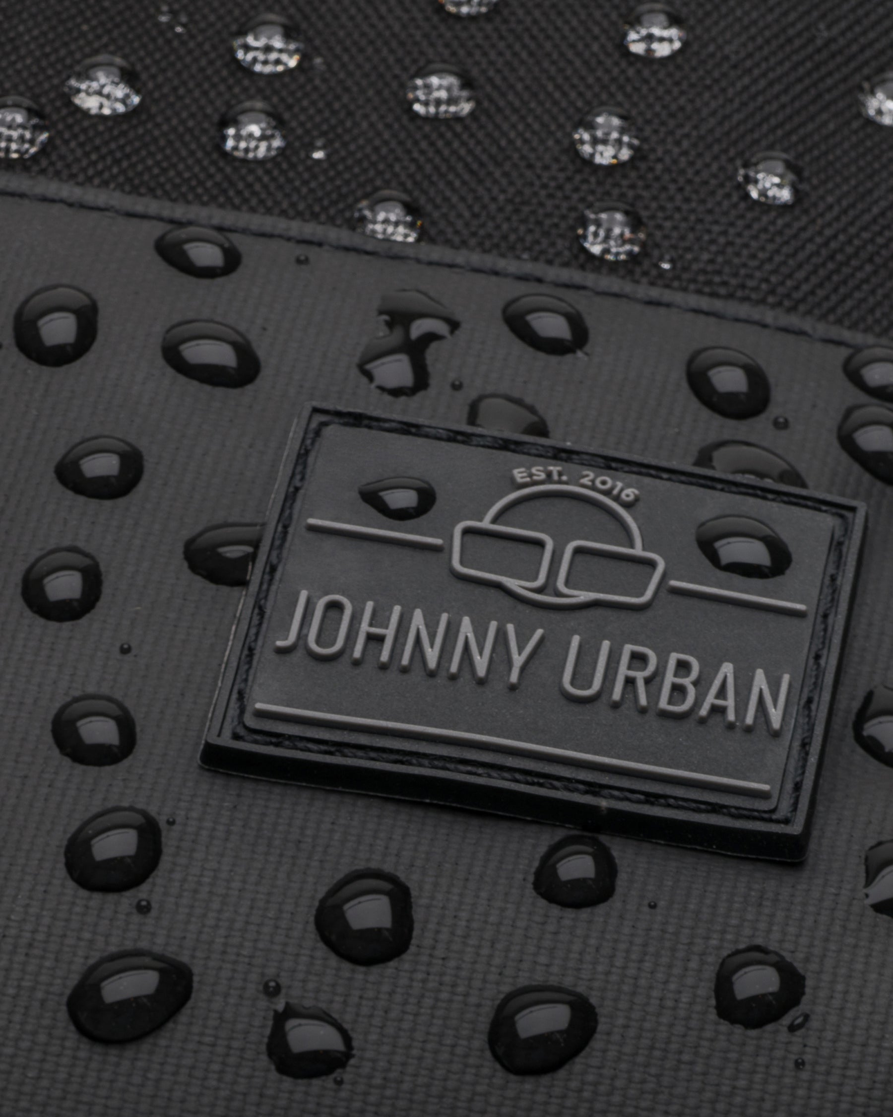 Nahaufnahme des wasserdichten schwarzen Rucksacks Mika von Johnny Urban mit Wassertropfen auf der Oberfläche und Markenlogo